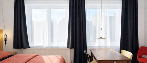A hotel room with a wooden floor, a bed with a pink bedspread, a wooden table with chairs, and a window with black curtains.