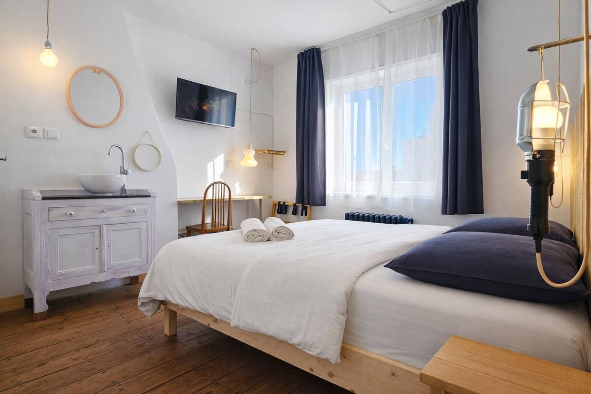 A bedroom with a wooden bed, white bedding, a bedside table, a mirror, a TV, a window with curtains, and a radiator.