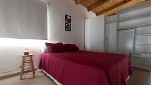Un dormitorio con una cama, un taburete de madera, un armario blanco y una ventana.