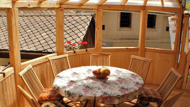 Een houten prieel met een ronde tafel en stoelen, een bloemen tafelkleed en een schaal fruit.