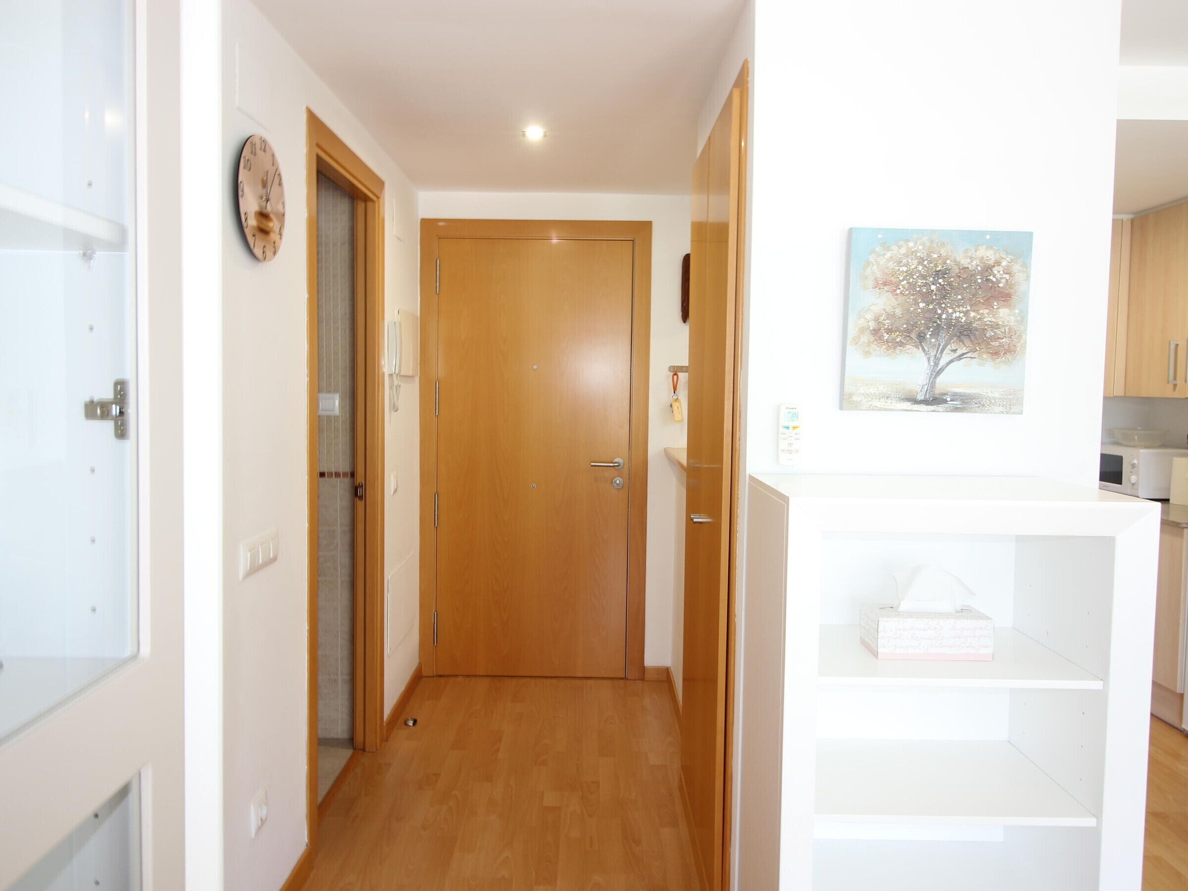 A hallway with a wooden door, a clock, a painting, and a white shelf with a box.