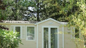 A small, single-story house with a beige exterior and white shutters. There is a door with a window above it, and a small porch area with white plastic chairs. - Mobile home with terrace - 6 guests (Montblanc)