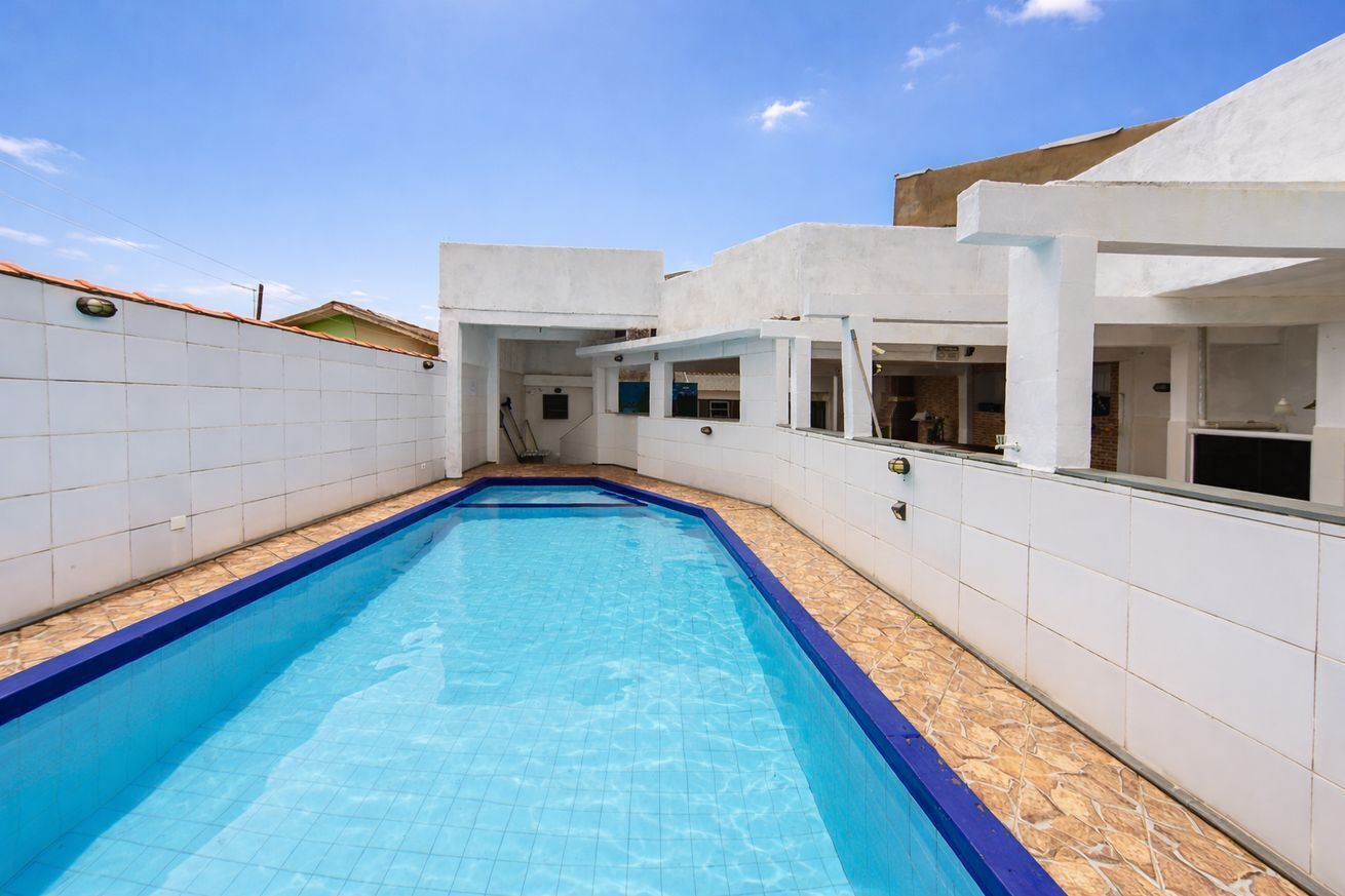 A swimming pool with a tiled deck, surrounded by white walls and a covered area with windows.