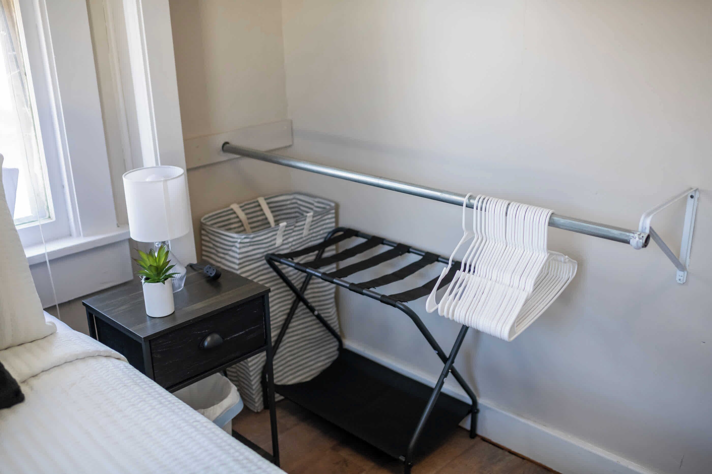 A bedroom with a bed, a bedside table with a lamp, a small plant, a black metal drying rack, and a striped basket.