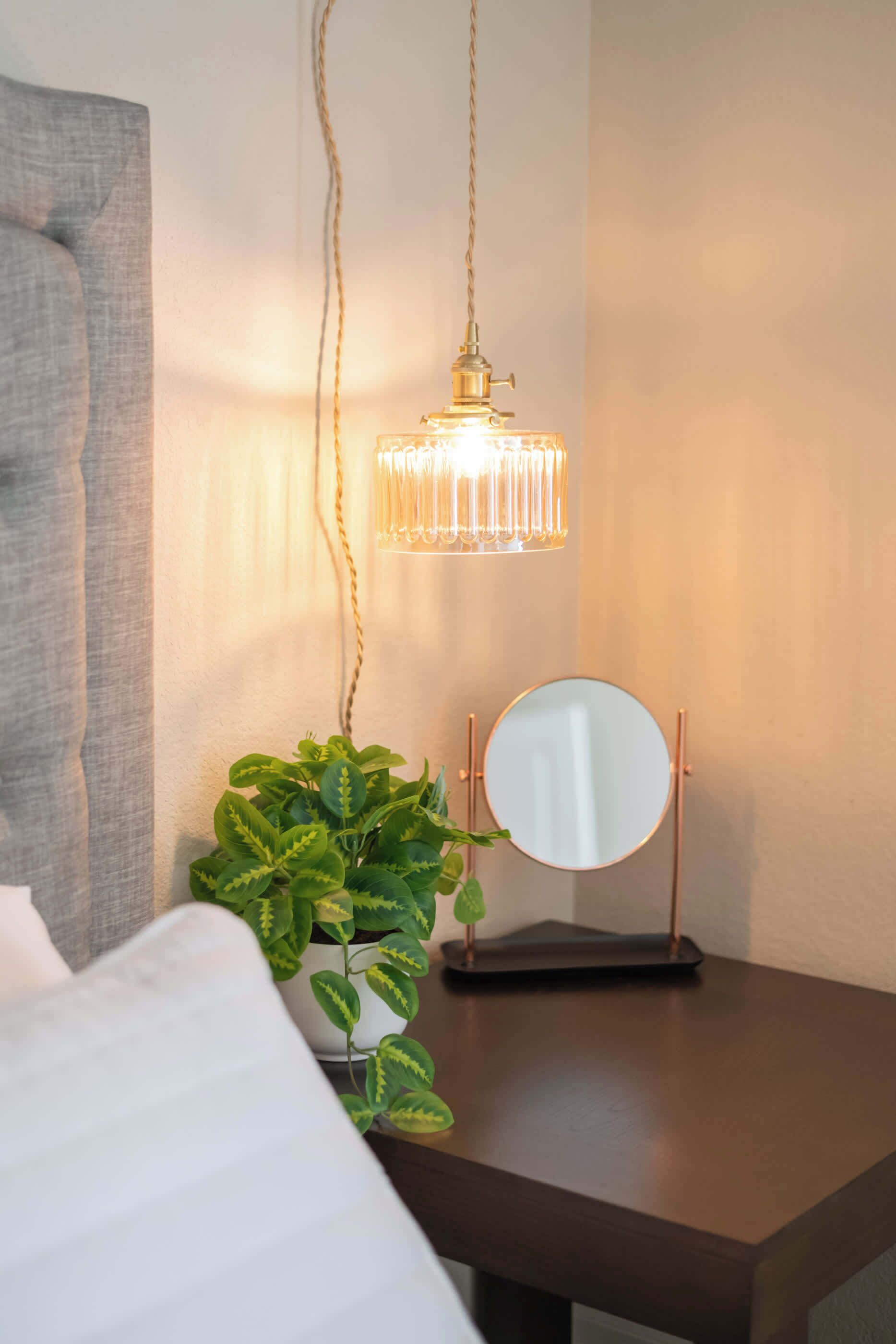 A neatly arranged bedroom with a bedside table featuring a round mirror, a potted plant, and a hanging light fixture.