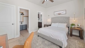 Une chambre à coucher comprenant un lit, un ventilateur de plafond, une table de chevet avec une lampe, une chaise et une porte donnant sur une salle de bain.