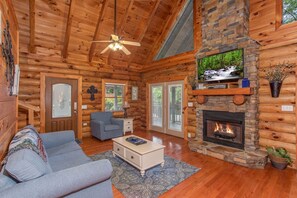 L’intérieur d’une cabane en rondins est agrémenté d’une cheminée en pierre, d’un téléviseur fixé au-dessus et d’un coin salon confortable avec un canapé et une table basse.