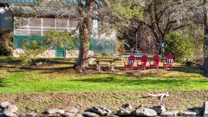 Un cadre pittoresque au bord de la rivière, avec une rangée de chaises Adirondack rouges, une table en pierre et un bâtiment surmonté d’une clôture verte en arrière-plan.