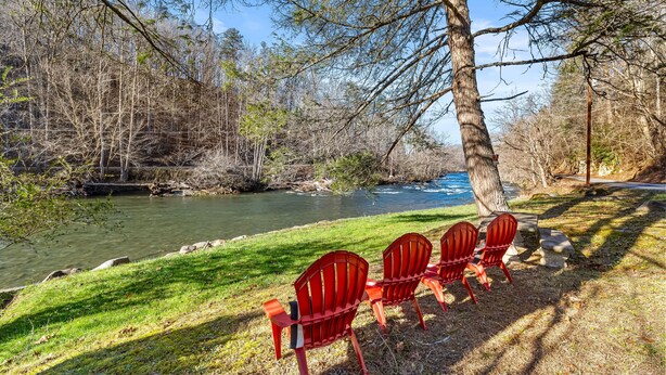 Une vue sur la rivière, avec quatre chaises Adirondack rouges disposées face à l’eau, entourées d’arbres et d’une zone herbeuse.