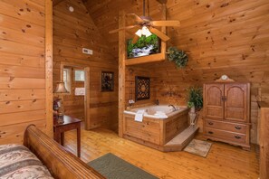 Interior de uma cabana de madeira com uma cama de madeira, um banheiro com banheira, um ventilador de teto e um armário de madeira.