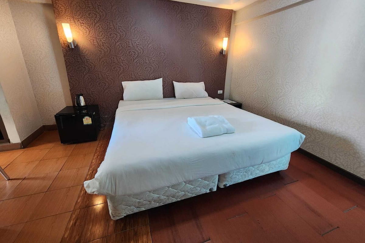 A neatly made bed with white linens and a folded towel, a bedside table with a lamp, and a patterned wall.