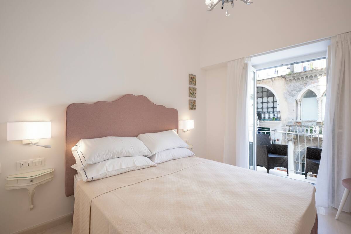 A bedroom with a large bed, bedside lamps, a nightstand, and a view of an outdoor balcony with chairs.
