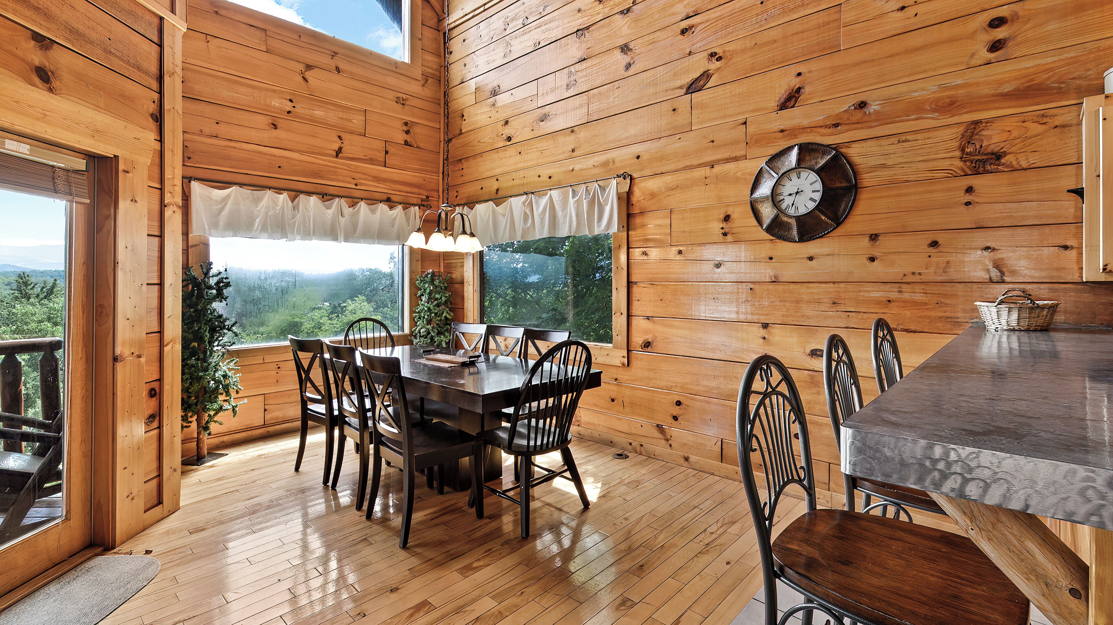 A wooden cabin with a dining area featuring a table and chairs, a metal clock on the wall, and a view of the outdoors through a window.