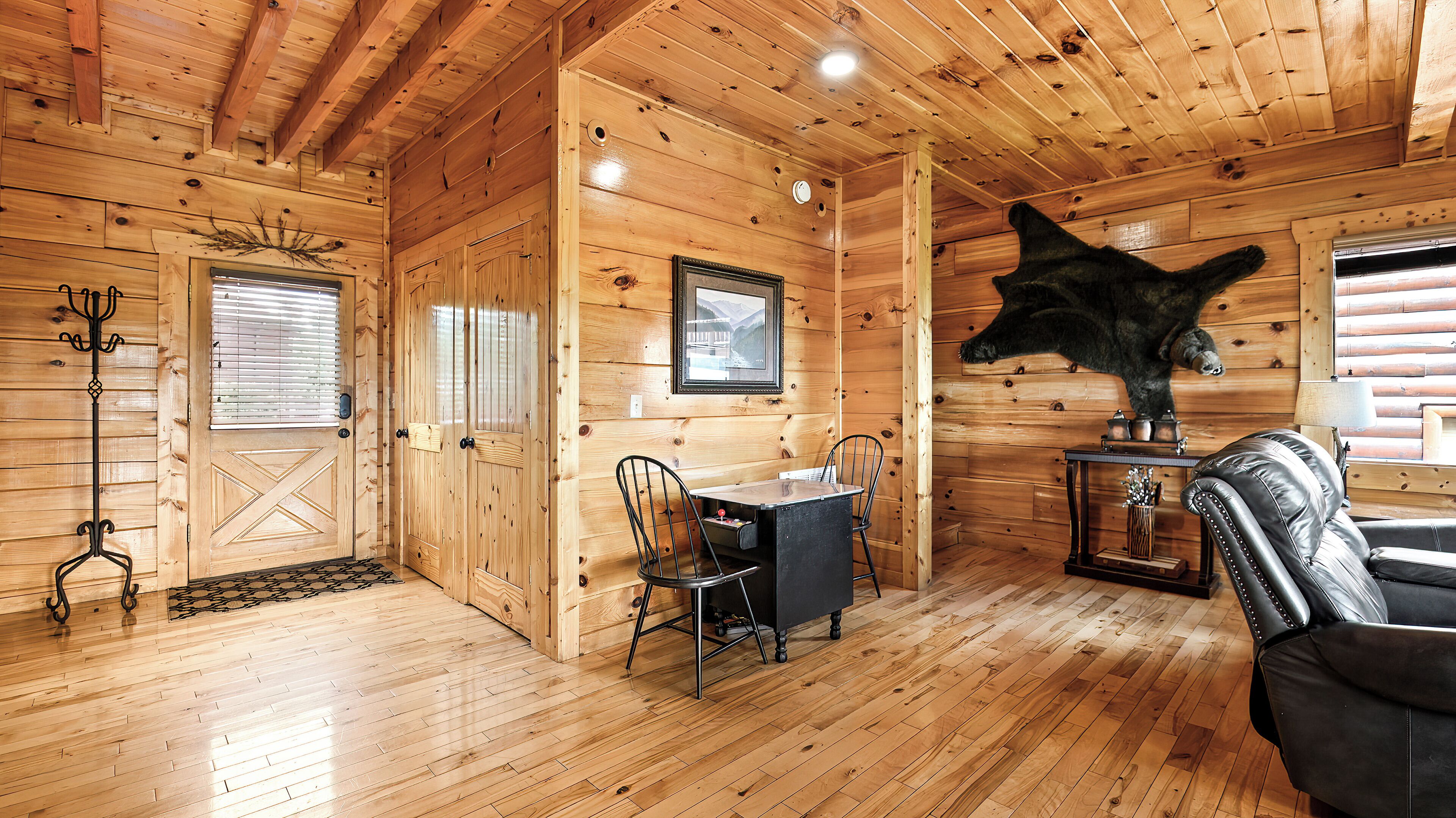 A wooden cabin interior with a dining table, chairs, a sofa, a mounted bear head, and a fireplace.