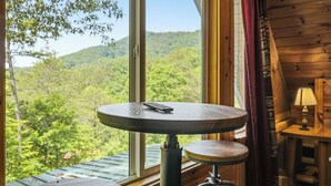 Ambiente in legno con un tavolo rotondo, una sedia e vista su un pendio boscoso attraverso una finestra.