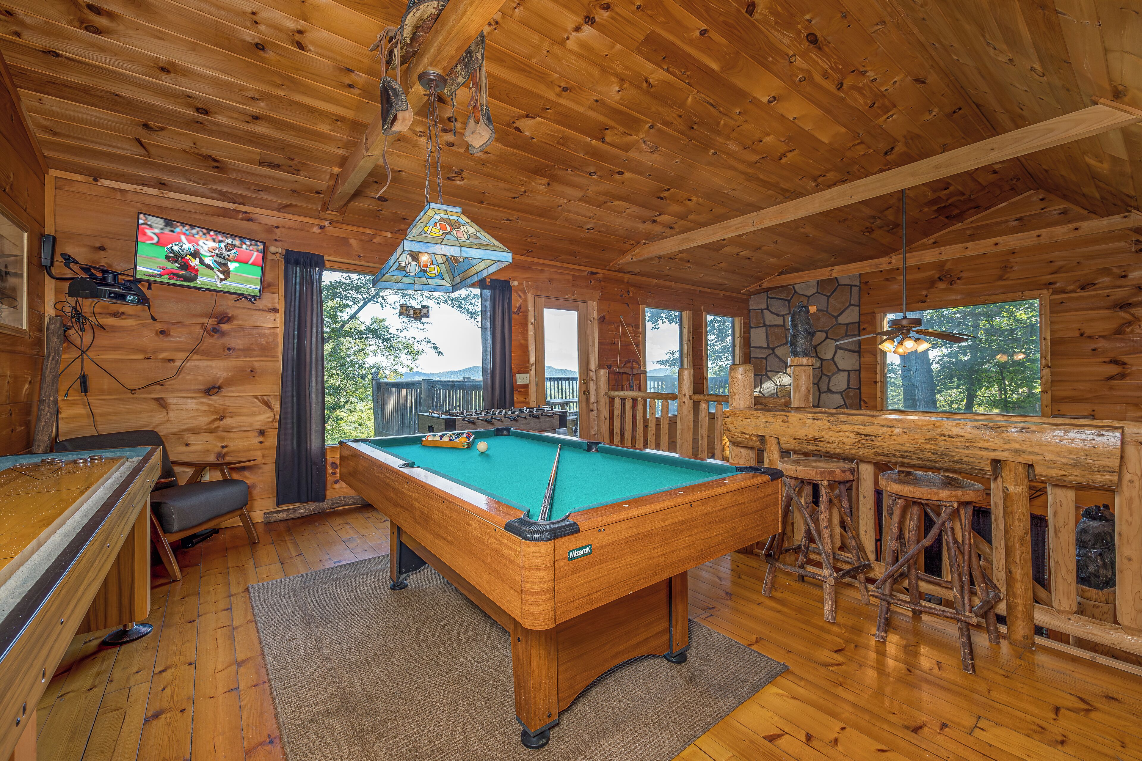 A wooden pool table in a cabin with a stone fireplace, wooden bar, and a television mounted on the wall.