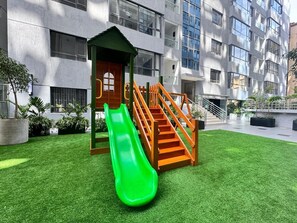 Ein Spielplatz mit einer grünen Rutsche und Treppen, umgeben von Gras und Topfpflanzen.