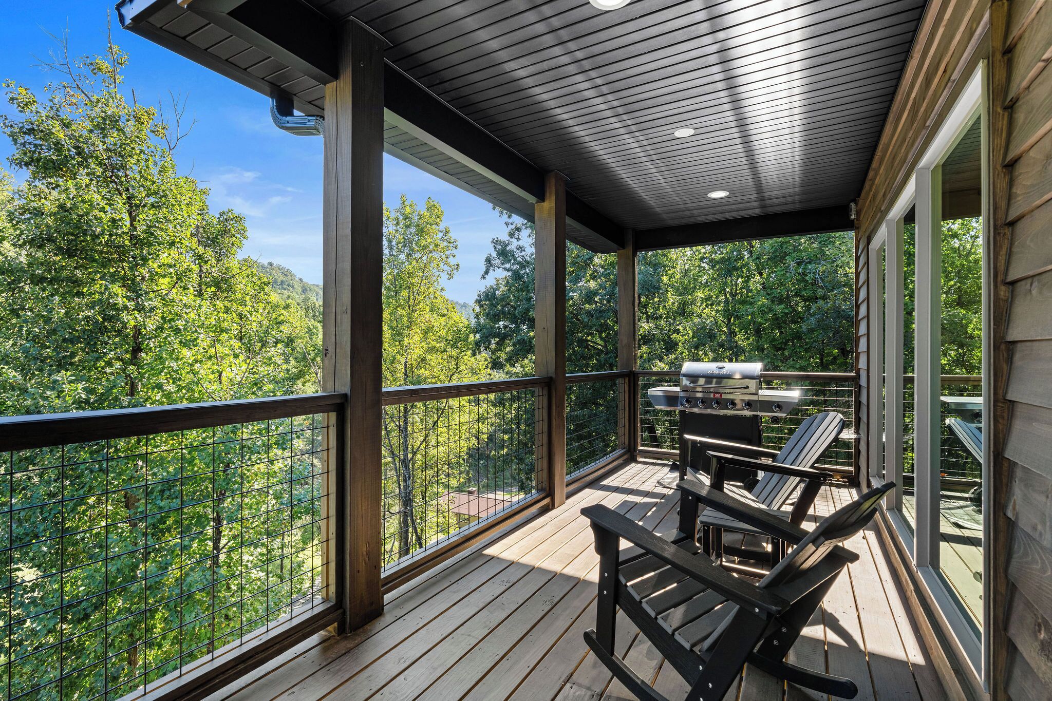 A wooden deck with a grill and chairs, overlooking a forested area.