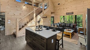 Ein großzügiges Blockhaus-Interieur mit einer zentralen Kochinsel, einer Treppe, einem Wohnbereich mit Sofa und Couchtisch und Blick ins Freie.