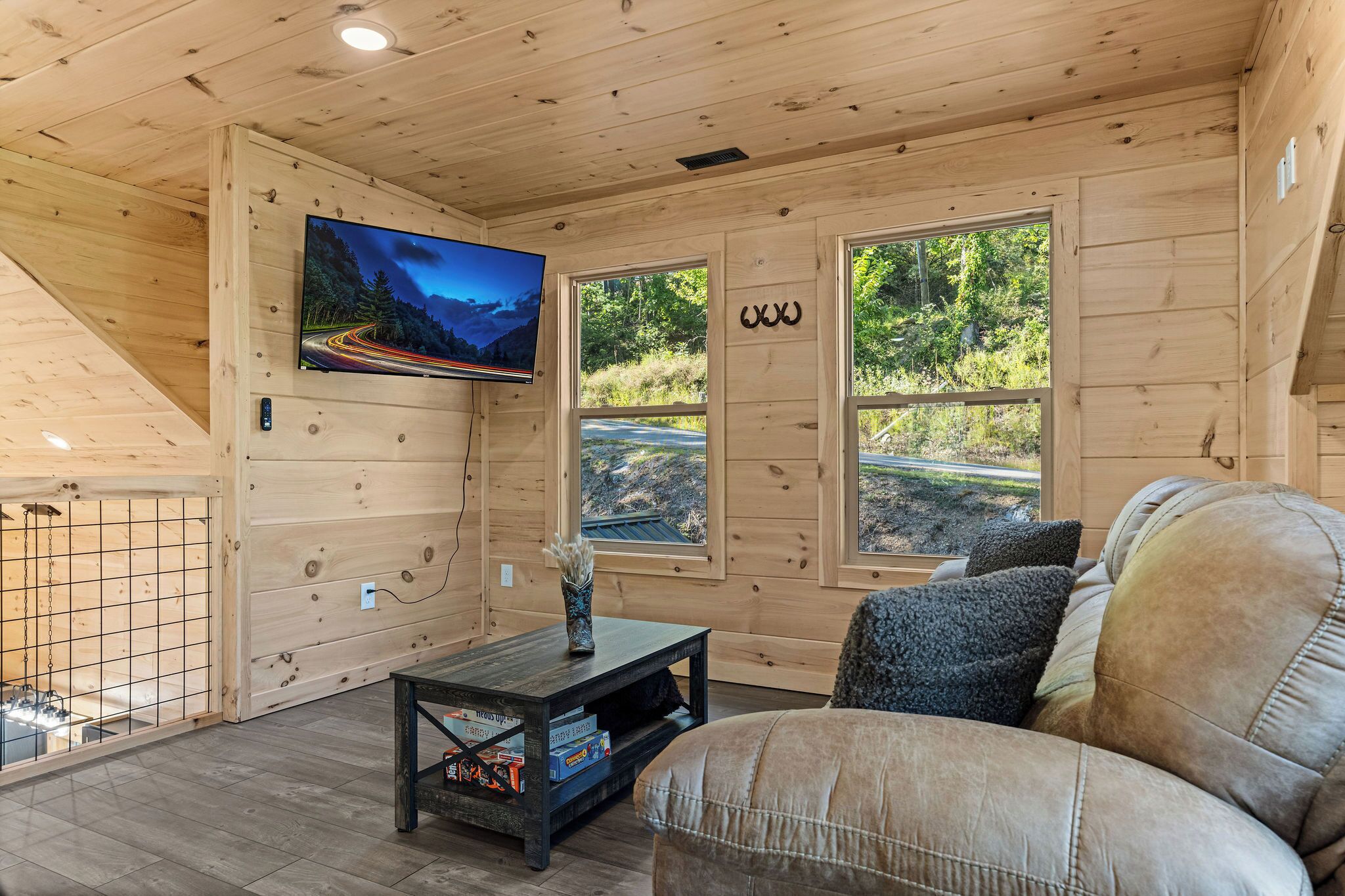 A wooden cabin interior with a TV mounted on the wall, a sofa, a small table, and large windows offering a view of the outdoors.