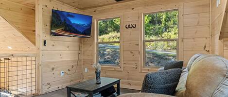 A wooden cabin interior with a TV mounted on the wall, a sofa, a small table, and large windows offering a view of the outdoors.