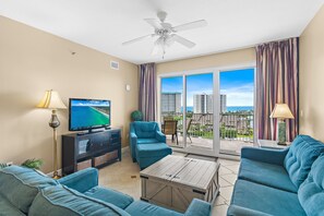 A living room with a TV, sofas, a ceiling fan, and a balcony with a view of buildings. - ⛱️Gulf Views | Walk to Beach | Sauna + 3 Pools | Ariel Dunes II 607🔆 (Miramar Beach)