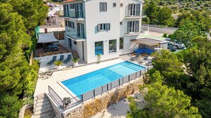 Ein modernes, mehrgeschossiges Haus mit einem großen Swimmingpool, umgeben von üppiger Vegetation und einer hügeligen Landschaft.