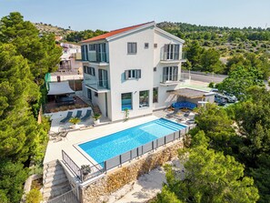 A modern multi-story house with a large swimming pool, surrounded by lush greenery and a hilly landscape. - Spacious Seafront Villa with Pool, Tennis, Gym & Mooring (Žaborić)