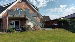 A two-story house with a brick facade, a balcony with railings, and a small garden area with potted plants and flowers. - Cozy vacation apartment with balcony. (Aschendorf)