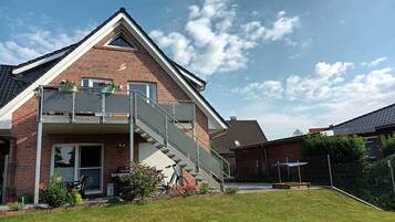 Een huis met twee verdiepingen, een gevel van baksteen, een balkon met leuningen, en een kleine tuin met potplanten en bloemen.