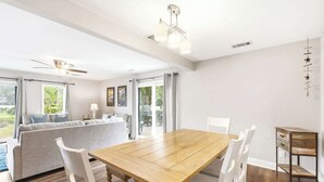 Een ruime eethoek met een houten tafel en witte stoelen, een bank met kussens, en een plafondventilator.