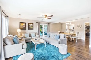Uma sala de estar espaçosa com um amplo sofá, uma mesa de centro e um tapete azul. Há uma área de jantar com mesa e cadeiras, e uma cozinha com fogão e forno.