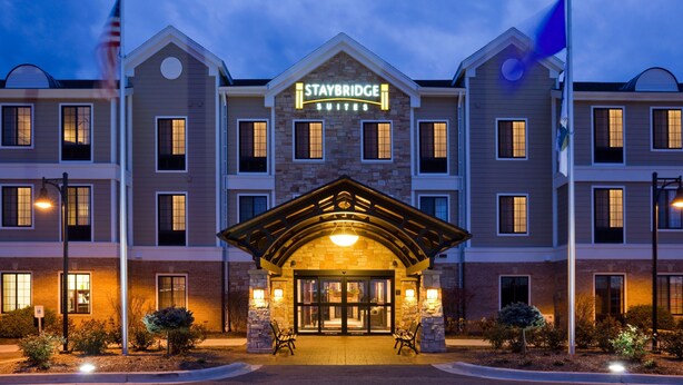 A Staybridge Suites hotel with a brick facade, a central entrance with a covered porch, and a flagpole with an American flag. - Accessible 1-BR Suite w/ Two Double Beds (Oconomowoc)