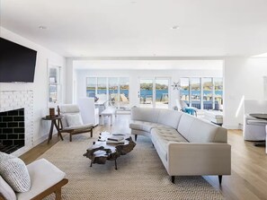 A spacious living room with a white sofa, a coffee table, and a fireplace. Large windows offer a view of the sea. - The Hamptons, Sag Harbor, Waterfront Escape (Sag Harbor)