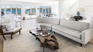 Een ruime woonkamer met een grote witte bank, een houten salontafel en uitzicht op zee door grote ramen.
