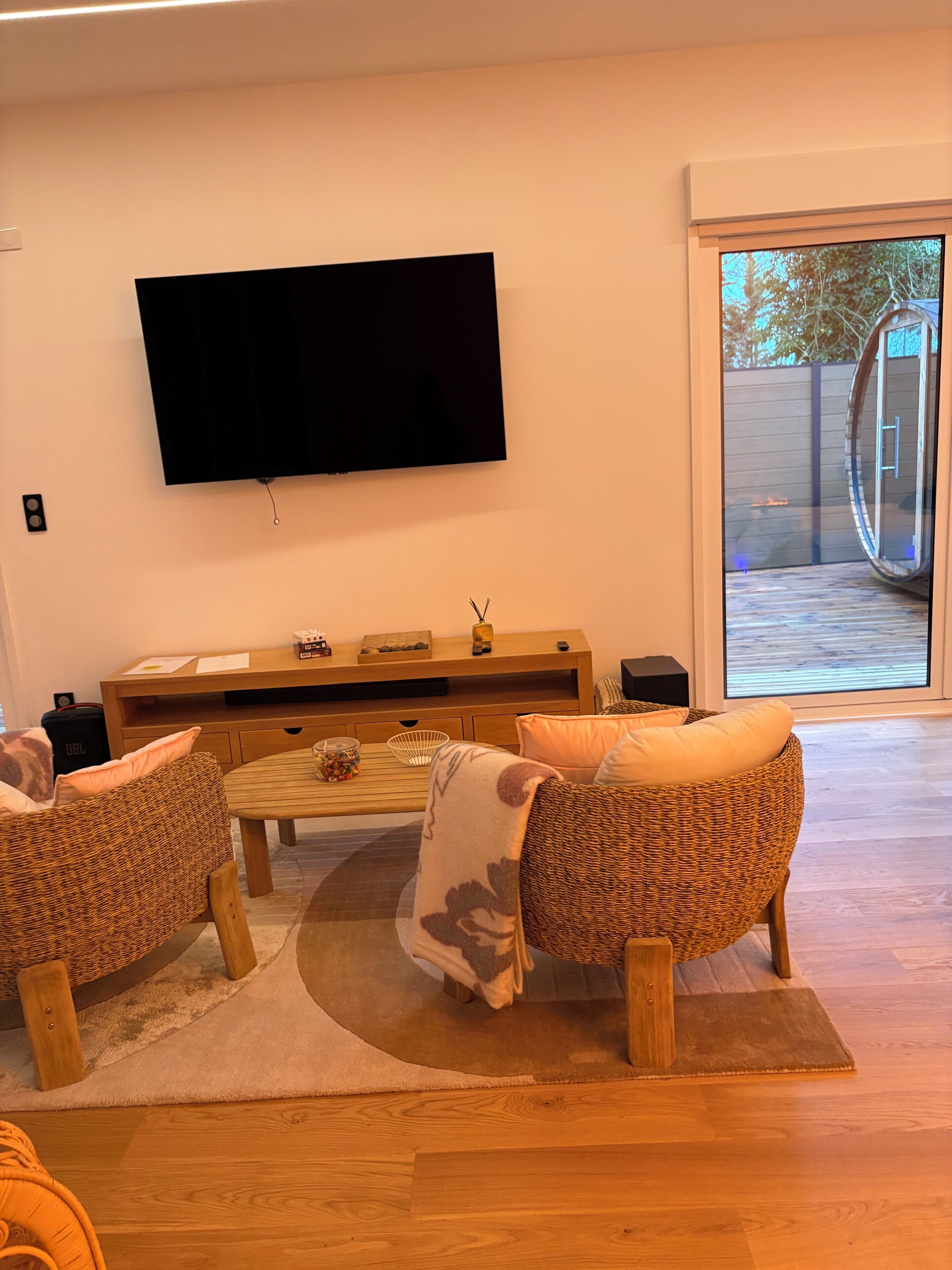 Een woonkamer met een flatscreen televisie aan de muur, een houten salontafel, rieten stoelen en een glazen deur naar buiten.