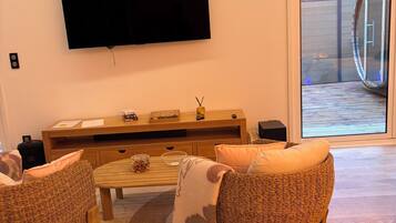 Een woonkamer met een flatscreen televisie aan de muur, een houten salontafel, rieten stoelen en een glazen deur naar buiten.