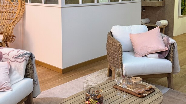 Een moderne woonkamer met een glazen scheidingswand, rieten meubels en een houten salontafel met decoratieve objecten.