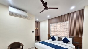 A hotel room with a bed, a desk, a chair, a ceiling fan, an air conditioning unit, and a window with blinds. - Hotel Gravity by De Tulip (Bhopal)