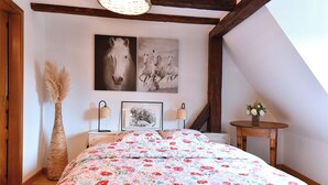 A bedroom with a floral bedspread, a large horse painting, a vase with flowers, and a lamp on a bedside table. - Gîte Vigne en fleur (Ribeauvillé)