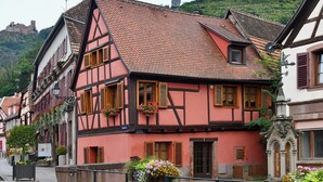 A traditional half-timbered building with a steeply pitched roof, a small balcony with flowers, and a stone wall in front. - Gîte Vigne en fleur (Ribeauvillé)