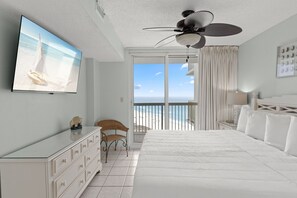 A bedroom with a large bed, a ceiling fan, a TV on the wall, a chair, and a view of the beach through the window. - Stunning Gulf Views, Multiple pools, Perfect blend of relaxation and convenience (Destin)