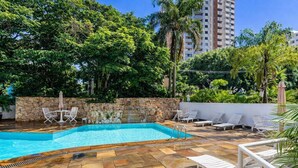 A swimming pool with stone walls and lounge chairs, surrounded by greenery and tall buildings. - Apto com Piscina e Garagem Perto do Ibirapuera (São Paulo)