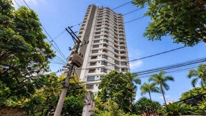 A tall, multi-story building with balconies, surrounded by lush greenery and palm trees. - Apto com Piscina e Garagem Perto do Ibirapuera (São Paulo)
