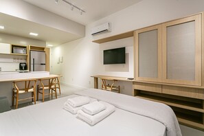 Quarto de hotel moderno com cama, escrivaninha, cadeira, televisão e kitchenette.