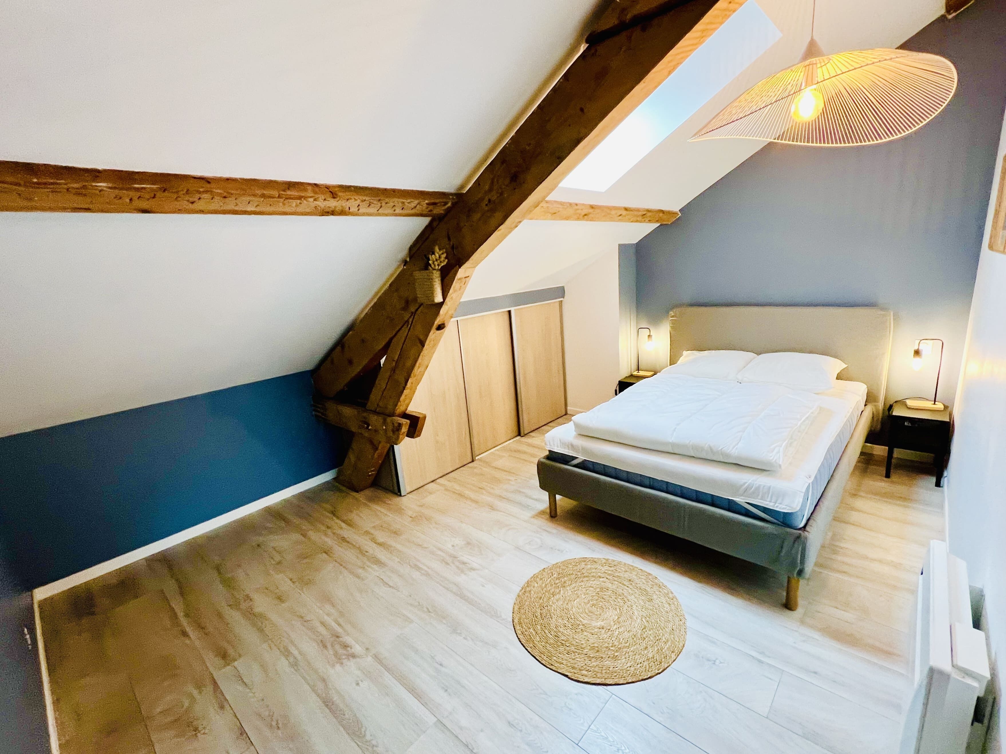 Une chambre avec un plafond en pente, des poutres en bois, un lit avec une literie blanche, un tapis tissé et une table de chevet avec une lampe.