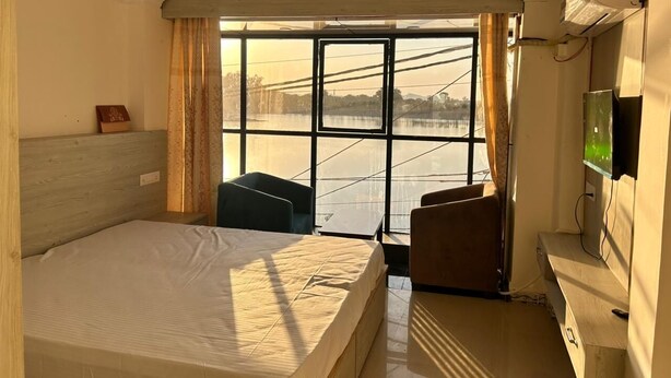 A hotel room with a bed, a desk, a chair, a TV, and a window with a view of a body of water. - Soni's Mansion Shivsagar (Khajuraho)