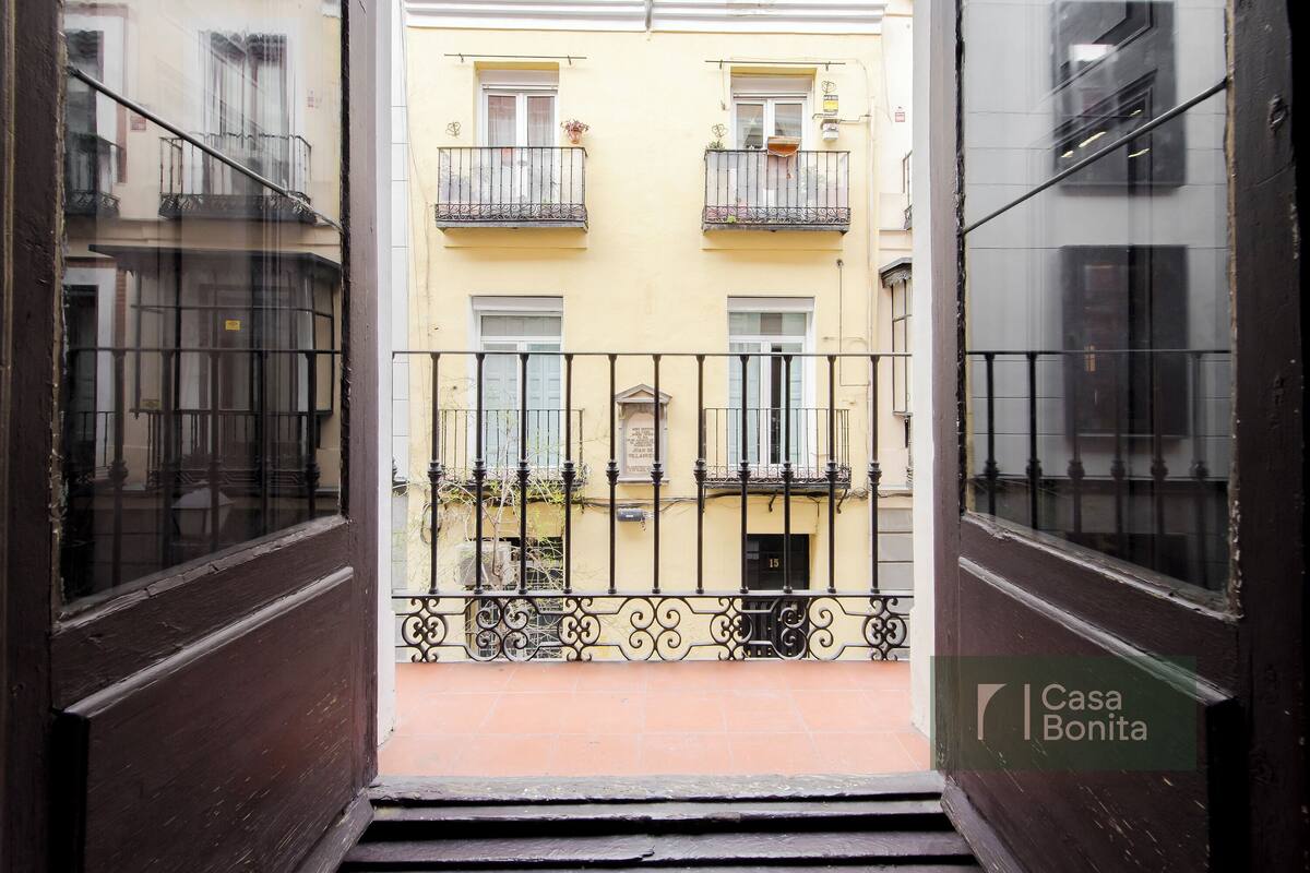 Vista a través de una puerta de madera hacia un edificio amarillo con balcones, piso de baldosas y barandilla de metal.