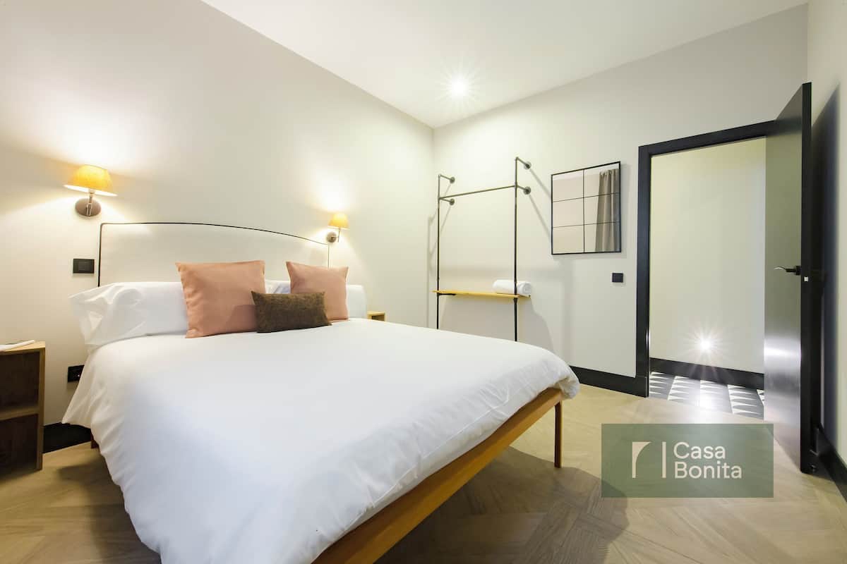 Un dormitorio moderno con una cama grande, lámparas de noche, un espejo y una puerta que da a otra habitación.