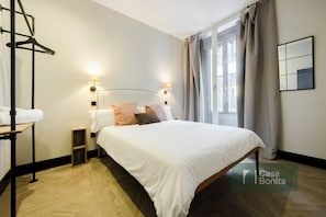 Un dormitorio ordenado con una cama, mesitas de noche, un perchero y una ventana con cortinas.